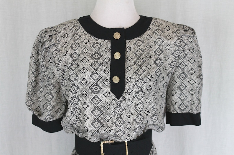 May include: A black and white patterned dress with a black collar and short sleeves. The dress has a black belt and gold buttons down the front.