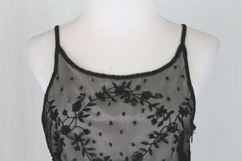 Pode incluir: Top preto com al&ccedil;as finas de espaguete com contas e design floral. O top &eacute; feito de tecido transparente e tem um decote redondo.