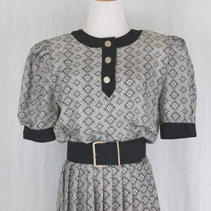 May include: A black and white patterned dress with a black belt and gold buttons. The dress has a round neckline and short sleeves.