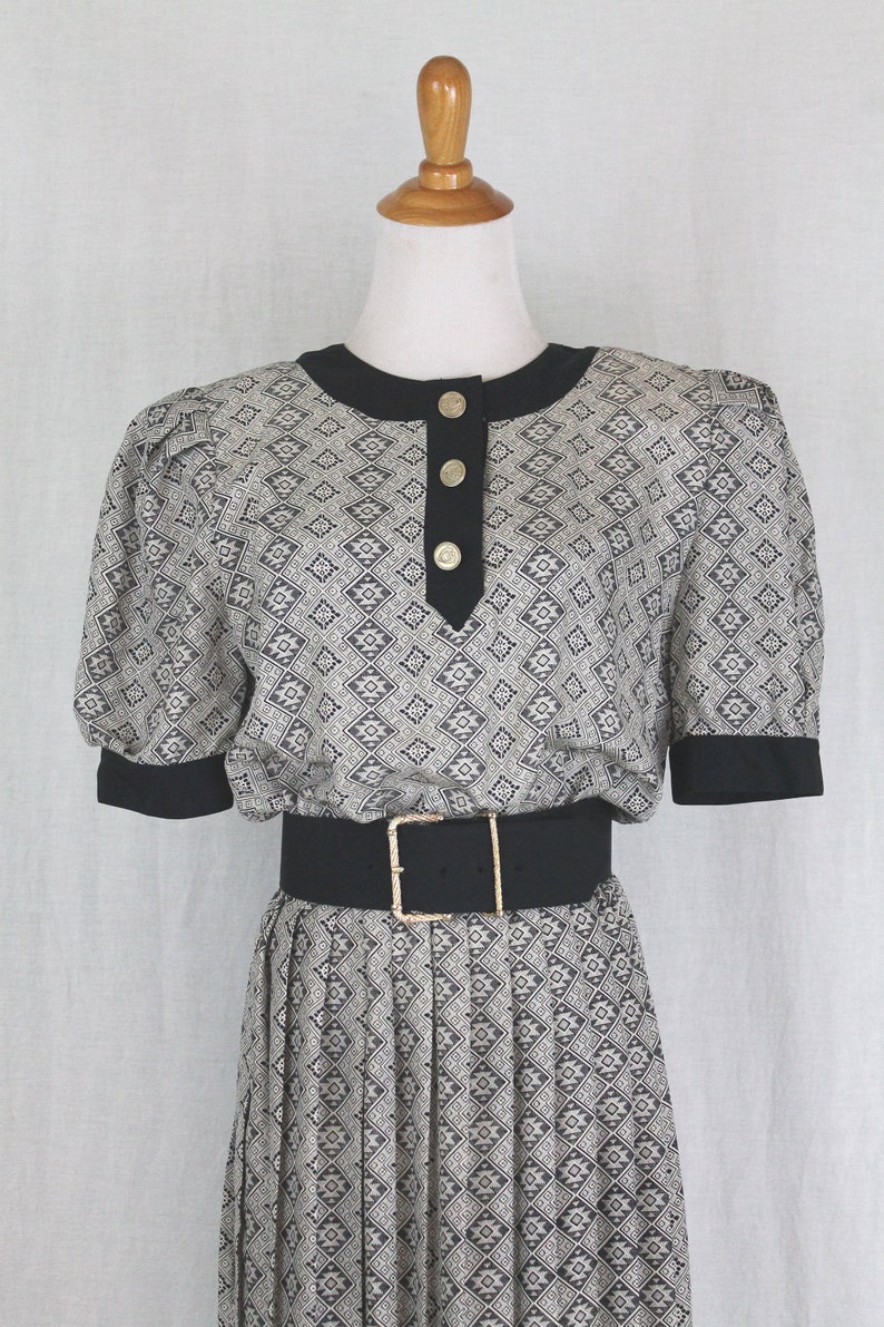 May include: A black and white patterned dress with a black belt and gold buttons. The dress has a round neckline and short sleeves.