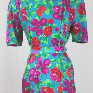 Puede incluir: Un vestido azul turquesa con estampado floral con flores rosas, rojas y verdes. El vestido tiene manga corta y un cintur&oacute;n.