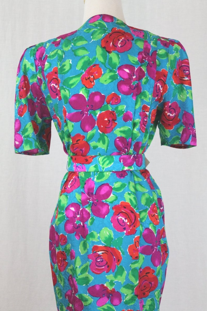 Puede incluir: Un vestido azul turquesa con estampado floral con flores rosas, rojas y verdes. El vestido tiene manga corta y un cintur&oacute;n.
