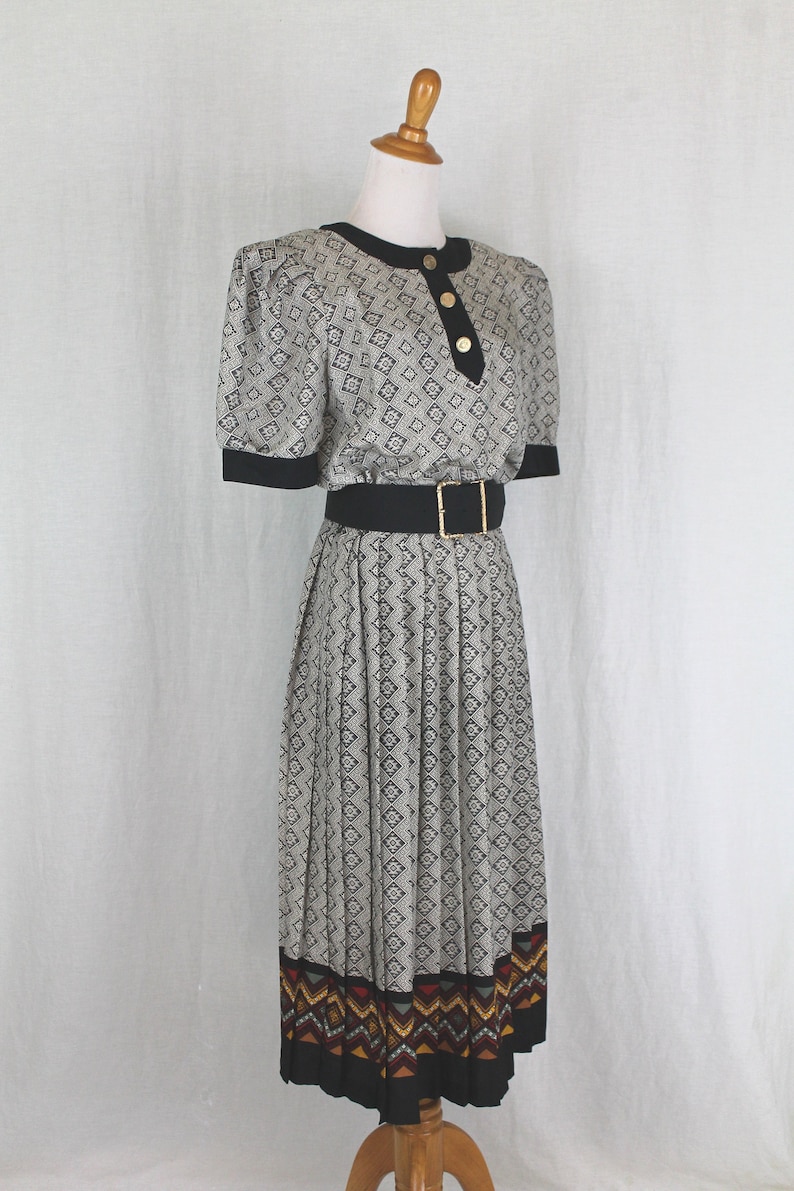 May include: A black and white patterned dress with a black belt and a colorful geometric trim at the hem. The dress has a fitted bodice and a pleated skirt.