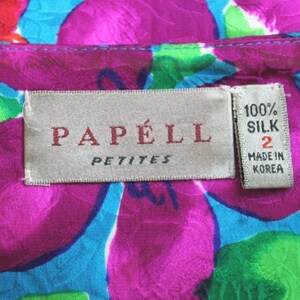 Puede incluir: Una etiqueta blanca con el texto "PAP&Eacute;LL PETITES" en letras rojas. Una etiqueta blanca m&aacute;s peque&ntilde;a con el texto "100% SILK 2 MADE IN KOREA" en letras negras est&aacute; unida a la etiqueta m&aacute;s grande. Las etiquetas est&aacute;n sobre una tela floral rosa, azul y verde.