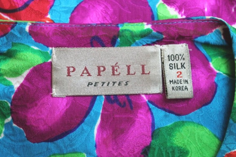 Puede incluir: Una etiqueta blanca con el texto "PAP&Eacute;LL PETITES" en letras rojas. Una etiqueta blanca m&aacute;s peque&ntilde;a con el texto "100% SILK 2 MADE IN KOREA" en letras negras est&aacute; unida a la etiqueta m&aacute;s grande. Las etiquetas est&aacute;n sobre una tela floral rosa, azul y verde.