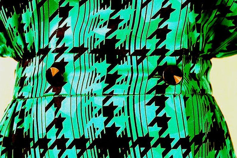 May include: A green and black patterned fabric with two black buttons. The fabric has a houndstooth pattern.