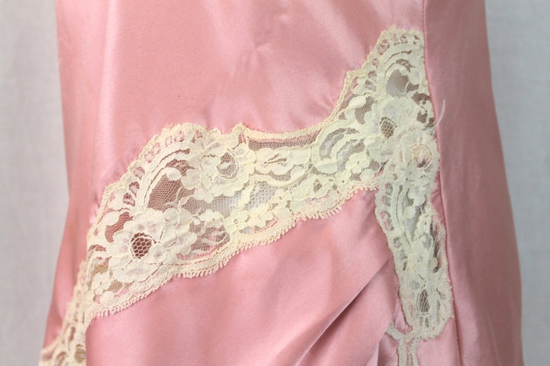 May include: A pink satin slip with a white lace trim. The lace trim is on the bottom edge of the slip and extends up the side.