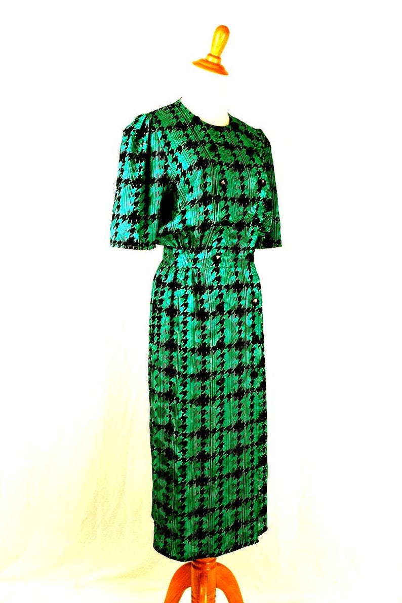 May include: A green and black houndstooth print dress with short sleeves and a button-up front. The dress is on a mannequin.