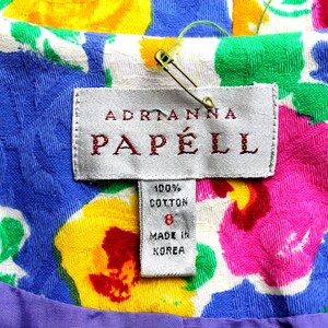 May include: A white dress tag with a floral pattern background. The tag reads "Adrianna Papell" in red lettering. The tag also includes a white label with the text "100% Cotton" and "Made in Korea".