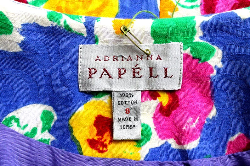May include: A white dress tag with a floral pattern background. The tag reads "Adrianna Papell" in red lettering. The tag also includes a white label with the text "100% Cotton" and "Made in Korea".