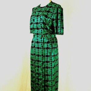 May include: A green and black houndstooth print dress with short sleeves and a fitted skirt. The dress is on a wooden mannequin.