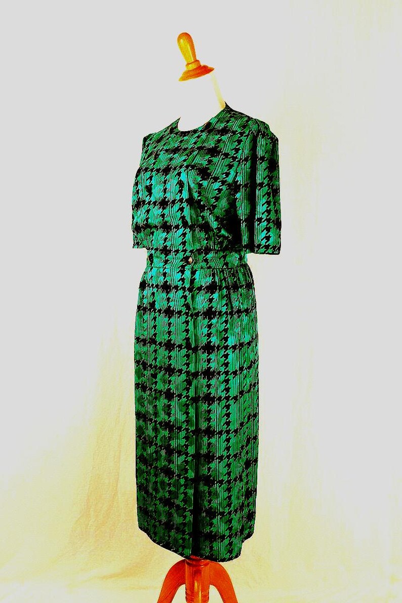 May include: A green and black houndstooth print dress with short sleeves and a fitted skirt. The dress is on a wooden mannequin.