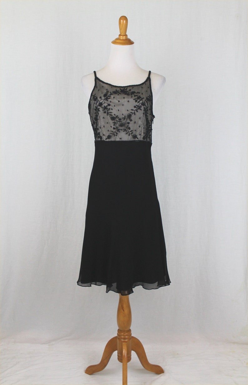 Pode incluir: Um vestido preto sem mangas com um corpete de renda transparente e uma saia preta. O vestido est&aacute; em um manequim de madeira.