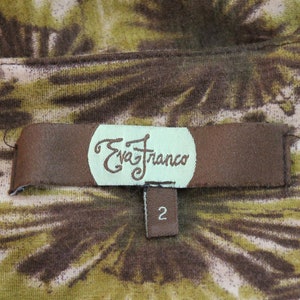 K&ouml;nnte beinhalten: Ein braunes Stofflabel mit dem Text "Eva Franco" in einem wei&szlig;en Kreis und der Nummer "2" auf einem kleineren braunen Etikett darunter. Das Label befindet sich auf einem braunen und gr&uuml;nen Stoff mit einem Blumenmuster.