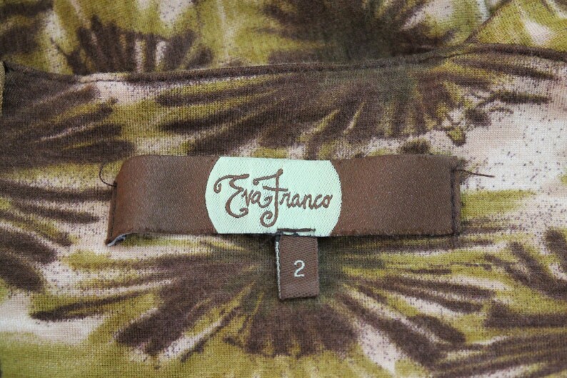 K&ouml;nnte beinhalten: Ein braunes Stofflabel mit dem Text "Eva Franco" in einem wei&szlig;en Kreis und der Nummer "2" auf einem kleineren braunen Etikett darunter. Das Label befindet sich auf einem braunen und gr&uuml;nen Stoff mit einem Blumenmuster.