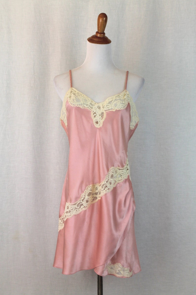 May include: A pink satin slip dress with delicate white lace trim. The lace is arranged in a diagonal pattern on the skirt and around the neckline.