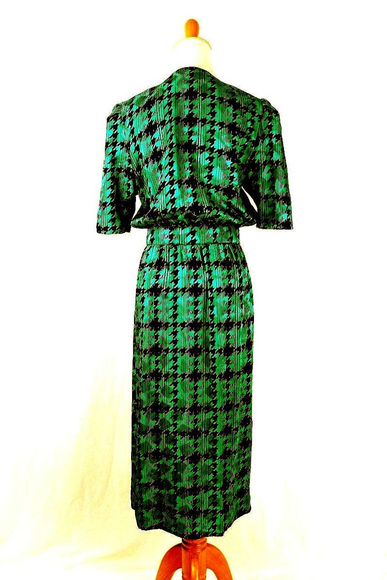 May include: A green and black houndstooth print dress with short sleeves and a fitted silhouette. The dress is on a mannequin and has a cinched waist.