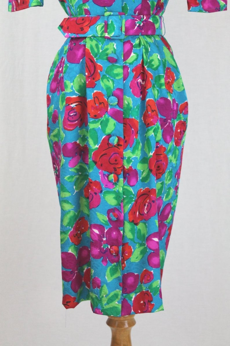 Puede incluir: Un vestido azul turquesa con estampado floral con flores rojas, rosas y verdes. El vestido tiene un frente abotonado y un cintur&oacute;n.
