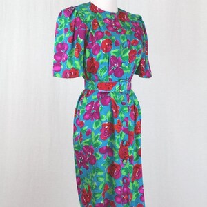 Puede incluir: Un vestido azul turquesa con estampado floral con flores rosas, rojas y verdes. El vestido tiene manga corta y un cintur&oacute;n.