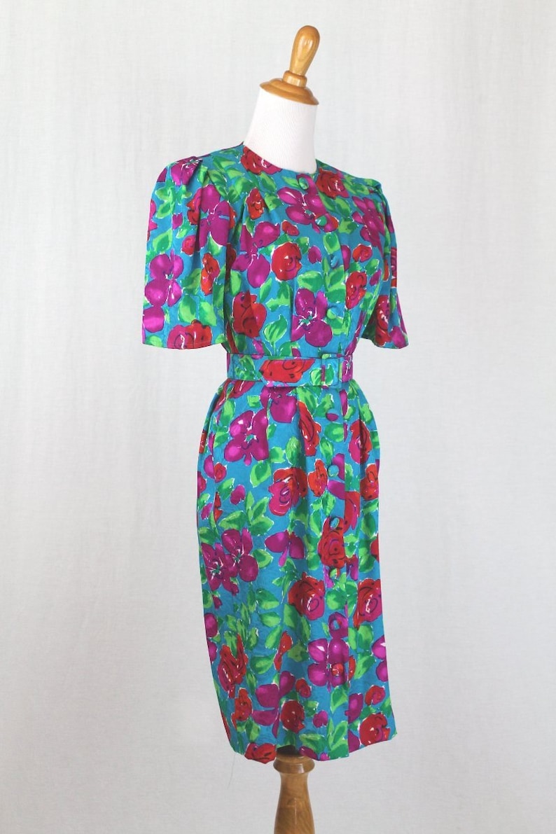 Puede incluir: Un vestido azul turquesa con estampado floral con flores rosas, rojas y verdes. El vestido tiene manga corta y un cintur&oacute;n.