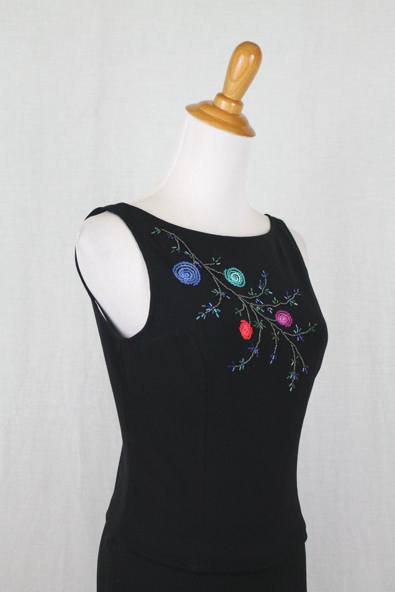 K&ouml;nnte beinhalten: Ein schwarzes, &auml;rmelloses Kleid mit einem floralen Stickmuster, das mit bunten Perlen verziert ist. Die Stickerei befindet sich auf der Vorderseite des Kleides und zeigt einen Zweig mit Bl&auml;ttern und Bl&uuml;ten.
