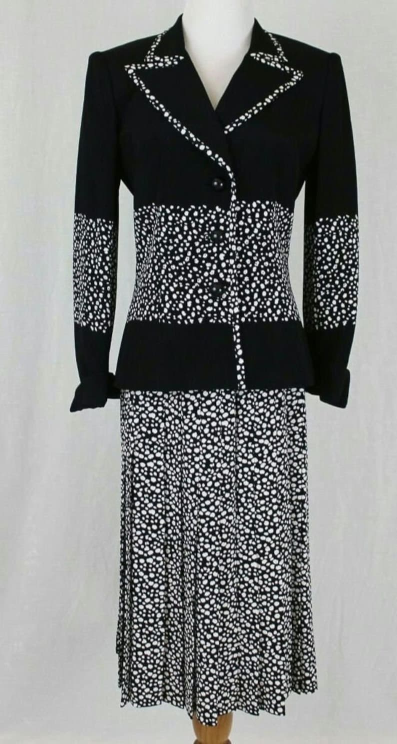 May include: A black and white polka dot skirt suit with a long-sleeved jacket and a skirt that falls to the knee. The jacket has a notched collar and a single button closure.