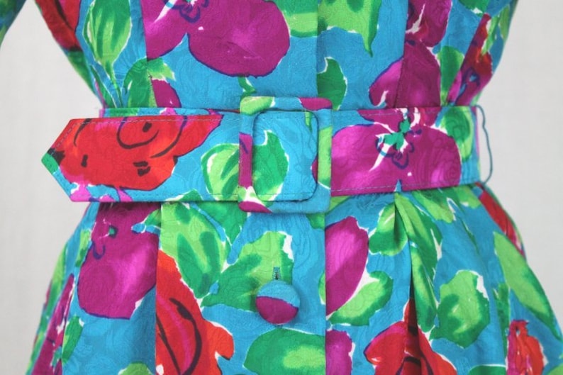 Puede incluir: Un vestido azul con estampado floral en tonos rosa, rojo y verde. El vestido tiene un cintur&oacute;n con una hebilla plateada.