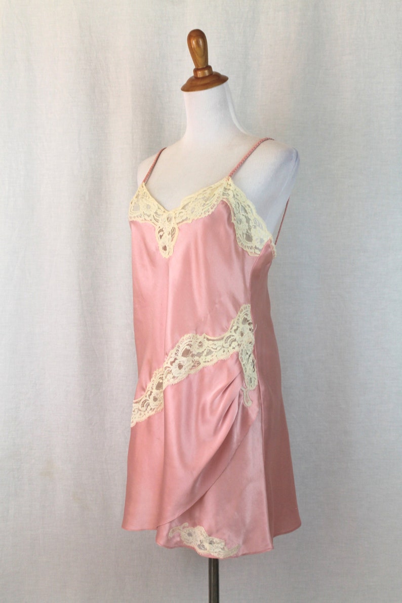 May include: A pink satin slip dress with delicate white lace trim. The dress has a V-neckline and adjustable straps.