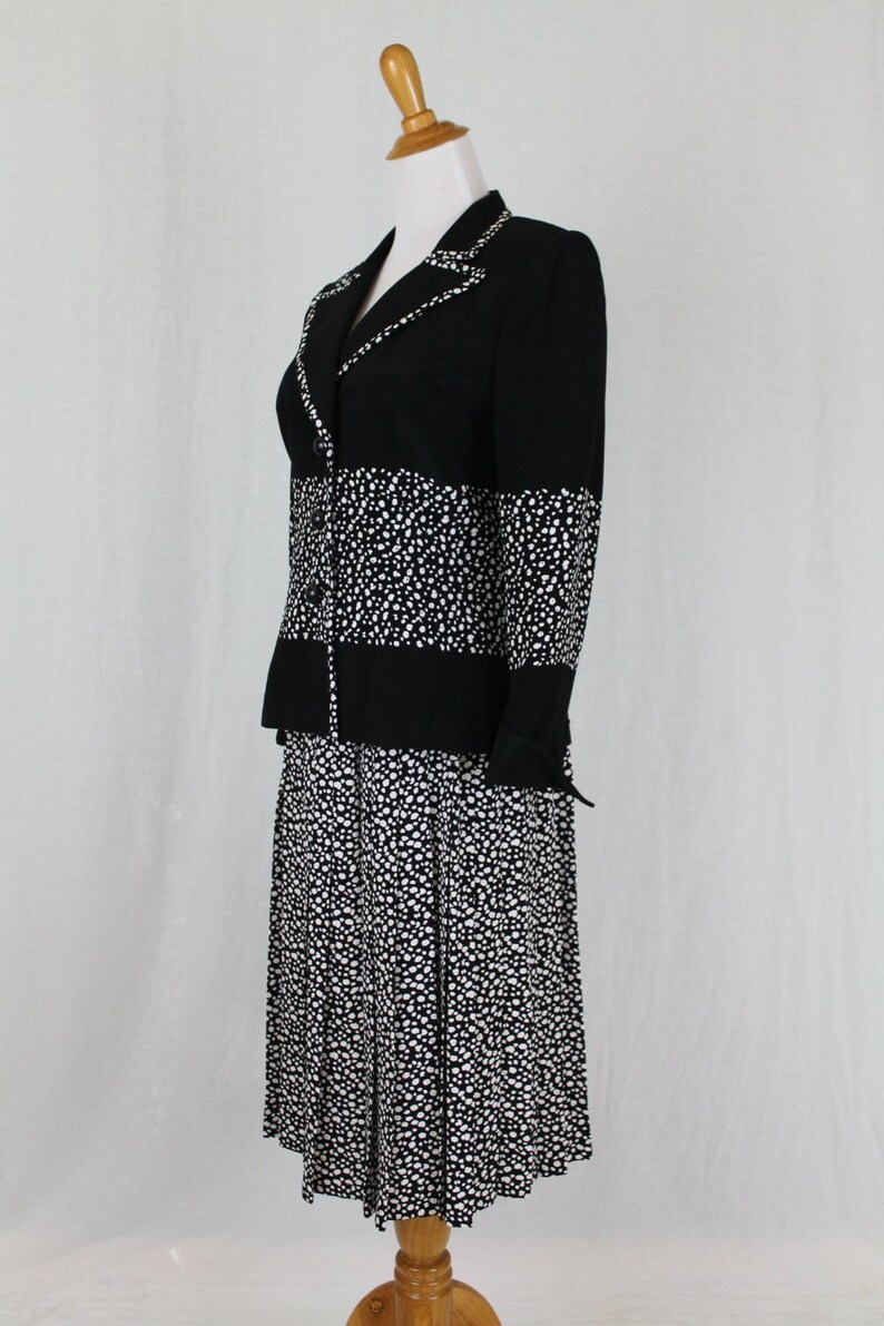 May include: A black and white polka dot skirt suit with a pleated skirt. The jacket has a black collar and a black band across the middle.