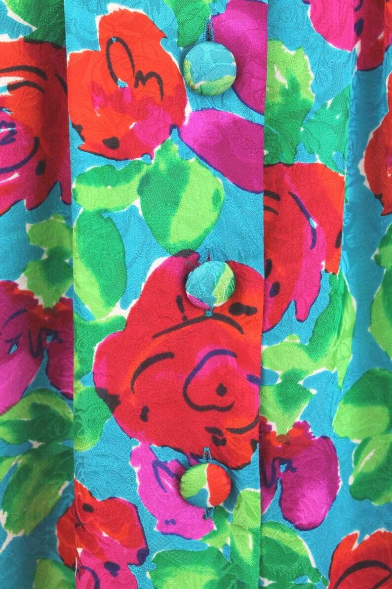 Puede incluir: Primer plano de una tela azul turquesa con un patr&oacute;n floral en tonos rojos, rosas, verdes y blancos. Tres botones azul turquesa est&aacute;n cosidos en la parte delantera de la tela.