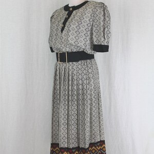 May include: A black and white patterned dress with a black belt and a colorful geometric border. The dress has a short sleeve and a pleated skirt.