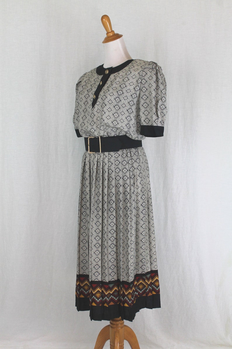 May include: A black and white patterned dress with a black belt and a colorful geometric border. The dress has a short sleeve and a pleated skirt.