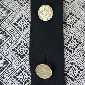 May include: A black fabric with a white and black geometric pattern. Two gold buttons with a crest design are attached to the fabric.