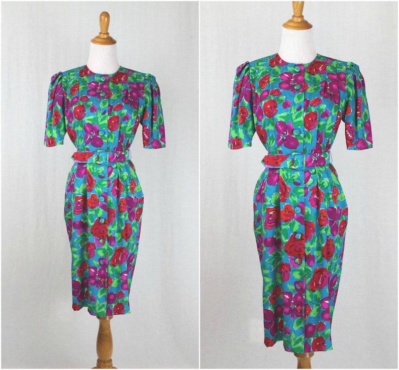 Puede incluir: Un vestido azul turquesa con estampado floral con grandes rosas rojas y rosas. El vestido tiene manga corta, cierre frontal con botones y un cintur&oacute;n a juego.