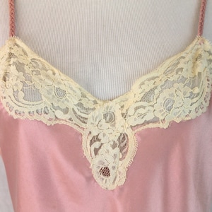 May include: A pink satin slip with delicate ivory lace trim around the neckline and a small lace detail at the center of the neckline.
