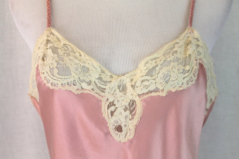 May include: A pink satin slip with delicate ivory lace trim around the neckline and a small lace detail at the center of the neckline.