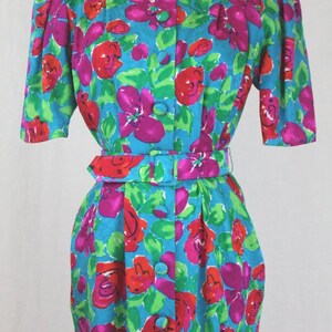 Puede incluir: Un vestido vintage floral con fondo turquesa y estampado floral a gran escala en tonos rosa, rojo y verde. El vestido tiene un frente abotonado, un cintur&oacute;n y mangas cortas.