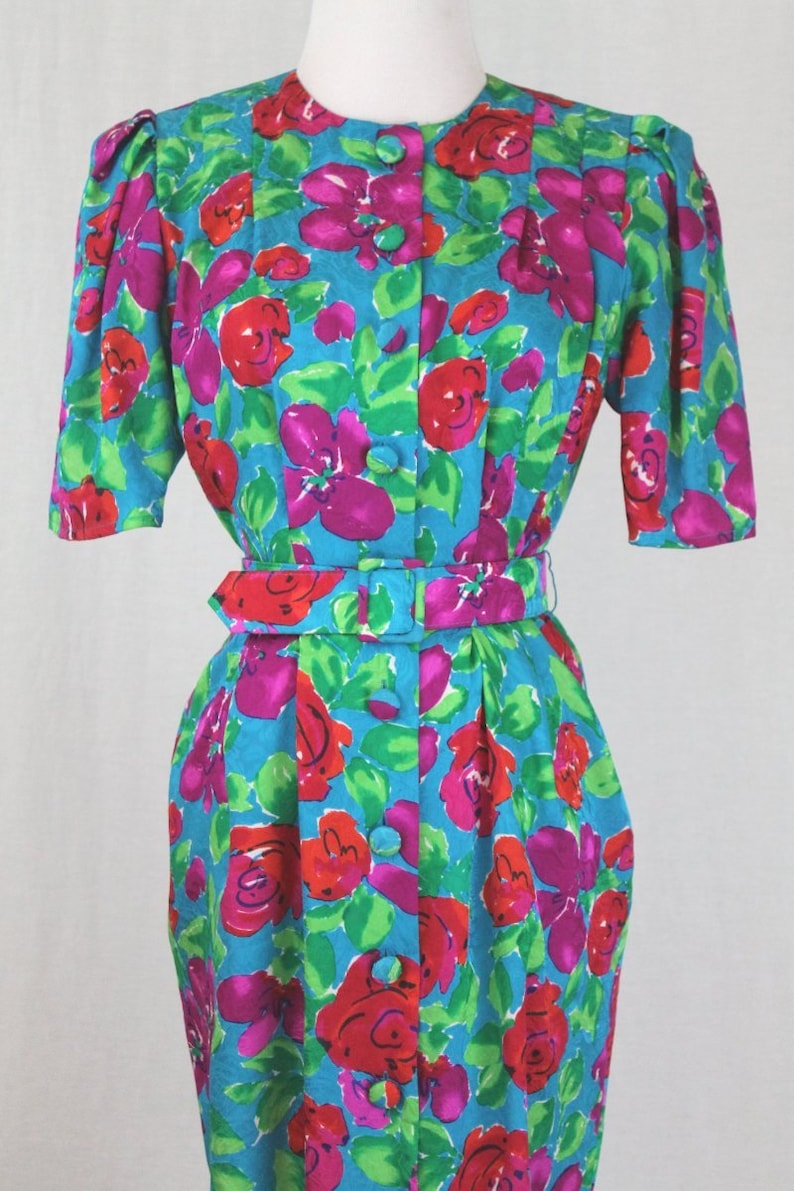 Puede incluir: Un vestido vintage floral con fondo turquesa y estampado floral a gran escala en tonos rosa, rojo y verde. El vestido tiene un frente abotonado, un cintur&oacute;n y mangas cortas.