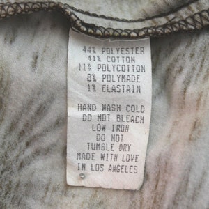 K&ouml;nnte beinhalten: Ein wei&szlig;es Etikett mit schwarzem Text, das die Stoffzusammensetzung eines Kleidungsst&uuml;cks beschreibt. Das Etikett lautet: 44% Polyester, 41% Baumwolle, 11% Polycotton, 8% Polymade, 1% Elasthan. Das Etikett enth&auml;lt auch Pflegehinweise: Handw&auml;sche kalt, nicht bleichen, B&uuml;geln bei niedriger Temperatur, nicht im Trockner trocknen. Das Etikett endet mit "Mit Liebe in Los Angeles hergestellt."