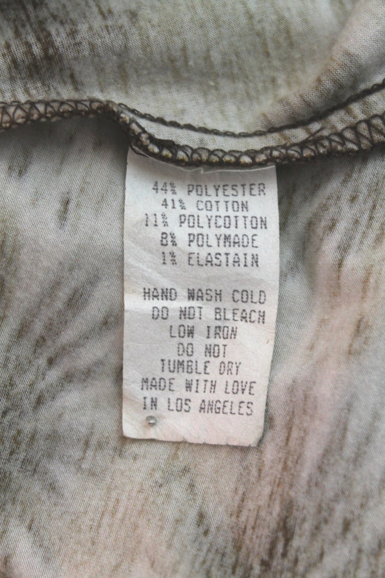 K&ouml;nnte beinhalten: Ein wei&szlig;es Etikett mit schwarzem Text, das die Stoffzusammensetzung eines Kleidungsst&uuml;cks beschreibt. Das Etikett lautet: 44% Polyester, 41% Baumwolle, 11% Polycotton, 8% Polymade, 1% Elasthan. Das Etikett enth&auml;lt auch Pflegehinweise: Handw&auml;sche kalt, nicht bleichen, B&uuml;geln bei niedriger Temperatur, nicht im Trockner trocknen. Das Etikett endet mit "Mit Liebe in Los Angeles hergestellt."