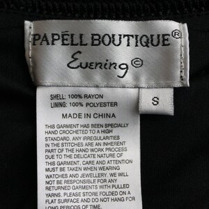 May include: White tag with black text that reads "PAPELL BOUTIQUE Evening" and "SHELL: 100% RAYON LINING: 100% POLYESTER MADE IN CHINA". The tag also includes a smaller white tag with the letter "S" on it. The tag is on a black fabric background.