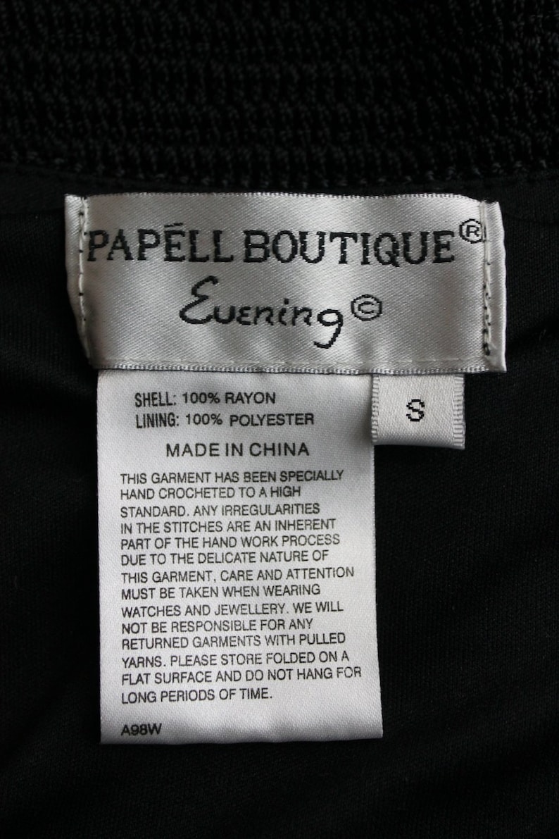 May include: White tag with black text that reads "PAPELL BOUTIQUE Evening" and "SHELL: 100% RAYON LINING: 100% POLYESTER MADE IN CHINA". The tag also includes a smaller white tag with the letter "S" on it. The tag is on a black fabric background.