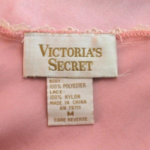 May include: A white tag with gold lettering that reads "VICTORIA'S SECRET" and lists the fabric content as 100% polyester for the body and 100% nylon for the lace. The tag also states "Made in China" and "RN 79711". The tag is attached to a pink garment and indicates a size "M" with instructions to "Care Reverse".