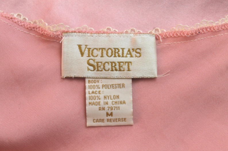 May include: A white tag with gold lettering that reads "VICTORIA'S SECRET" and lists the fabric content as 100% polyester for the body and 100% nylon for the lace. The tag also states "Made in China" and "RN 79711". The tag is attached to a pink garment and indicates a size "M" with instructions to "Care Reverse".