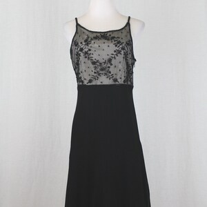 Pode incluir: Um vestido preto sem mangas com um corpete de renda preta transparente com um padr&atilde;o floral. O vestido tem um corpete justo e uma saia fluida.