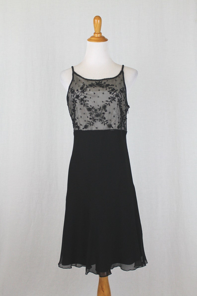 Pode incluir: Um vestido preto sem mangas com um corpete de renda preta transparente com um padr&atilde;o floral. O vestido tem um corpete justo e uma saia fluida.