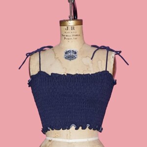 May include: A navy blue crop top with tie straps, displayed on a vintage dress form. The top has a smocked bodice and a ruffled edge. The dress form is cream-colored, with the number 12 and the year 1960 printed on it. The background is a solid pink.