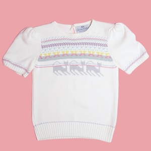 May include: Cream-colored short-sleeved sweater with a colorful geometric pattern across the chest. The pattern includes yellow, pink, and purple accents, and a row of gray animal silhouettes. The sweater has a ribbed hem and cuffs.