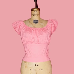 May include: A pink, off-the-shoulder blouse with short sleeves on a dress form. The dress form is labeled "Model 12 1960".