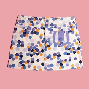 May include: A white mini skirt with a colourful polka dot pattern in shades of blue, orange, and purple. The skirt has a single pocket on the right side with a blue and white striped trim.